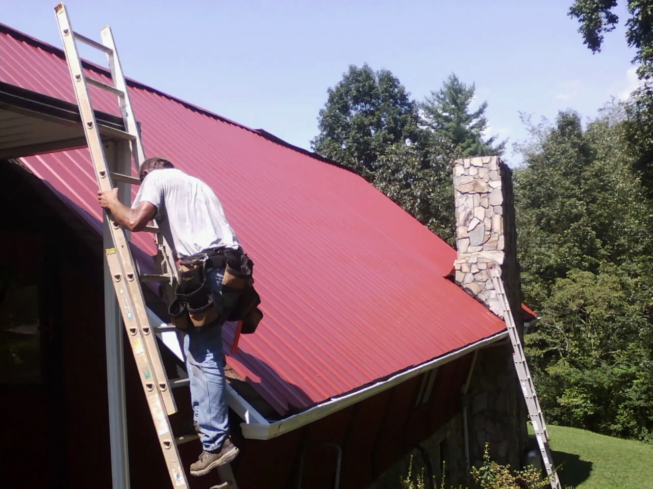 Roofer on a ladder beside a red metal roof and stone chimney — Soffit & Fascia Repair by Roofing Brothers in Council Bluffs